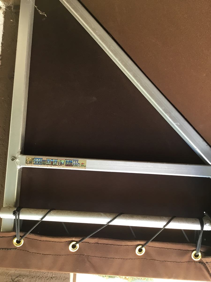 Brown residential awning with durable canvas material, reinforced by a sturdy aluminum frame and secured with black cords and grommets.