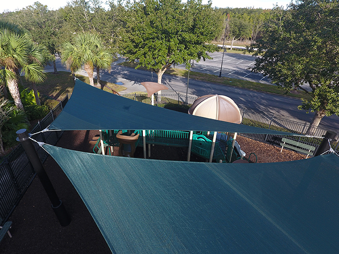 Green shade sails providing sun protection over a playground area, supported by sturdy black posts in a landscaped outdoor setting.